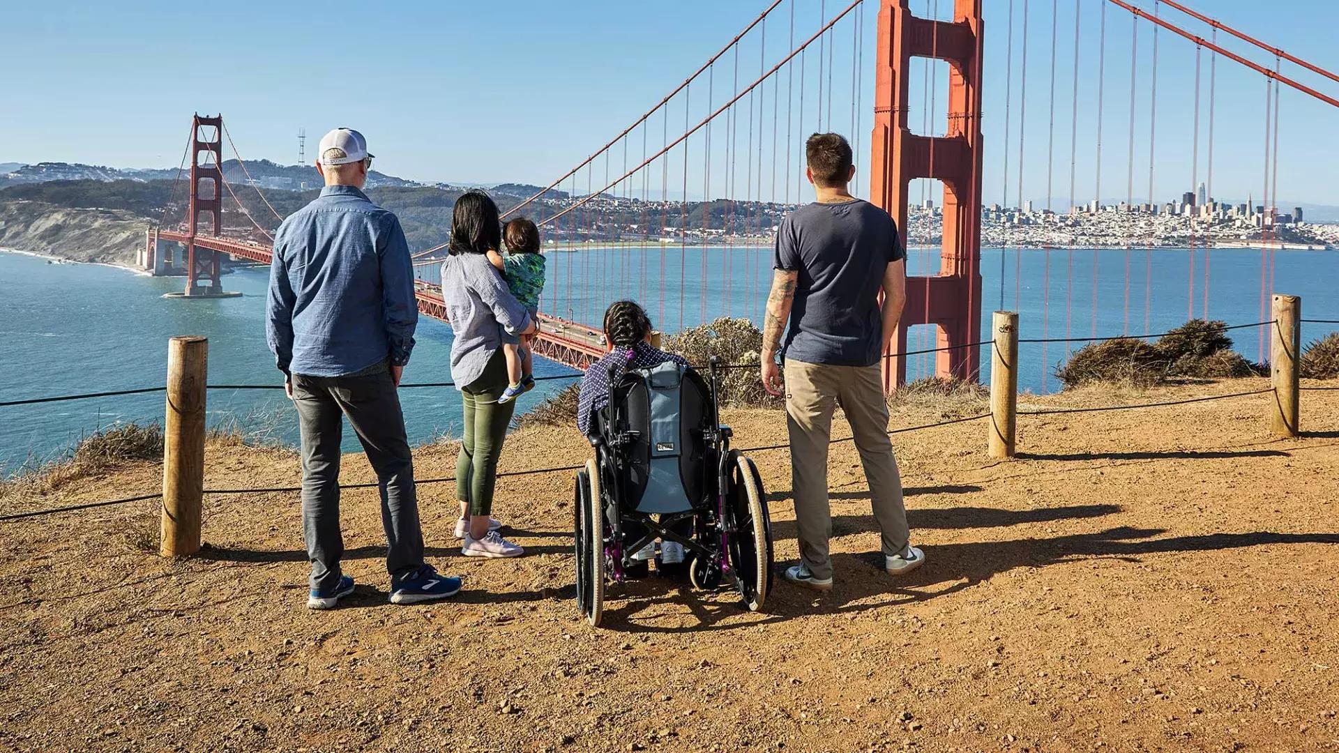 A group of people, including one person in a wheelchair, is seen from behind as they look at the Golden Gate Bridge from the Marin Headlands.