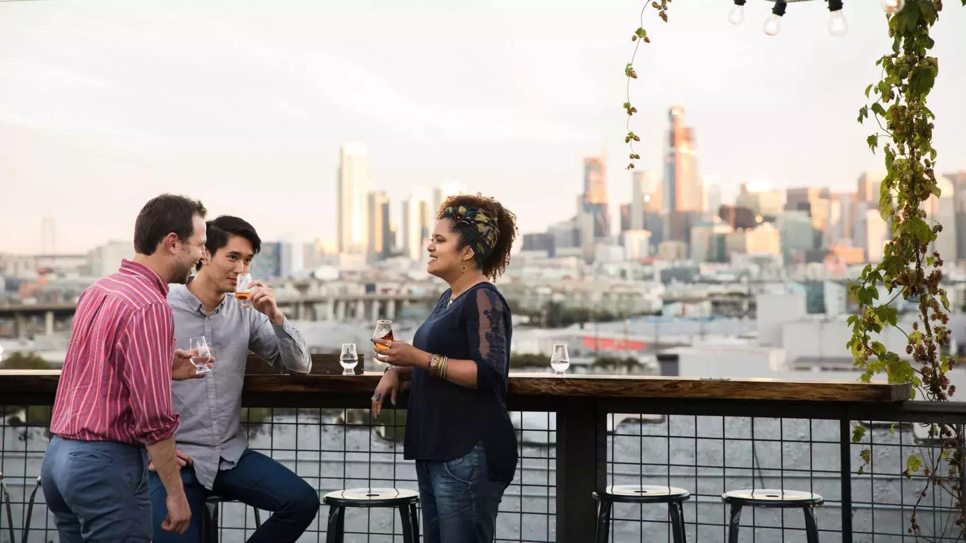 カリフォルニア州サンフランシスコにあるアンカー・ディスティリング社の屋上デッキにある屋外テーブルの周りに3人が集まっています。