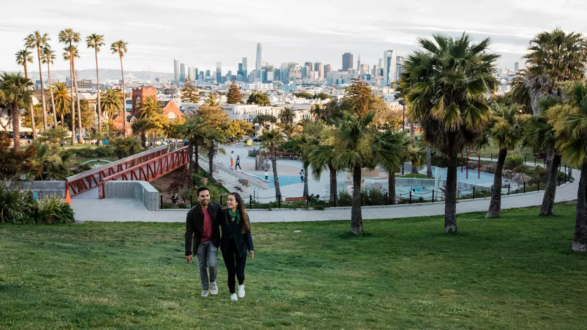 한 커플이 돌로레스 공원과 샌프란시스코 스카이라인을 뒤로하고 카메라를 향해 걸어가고 있습니다.