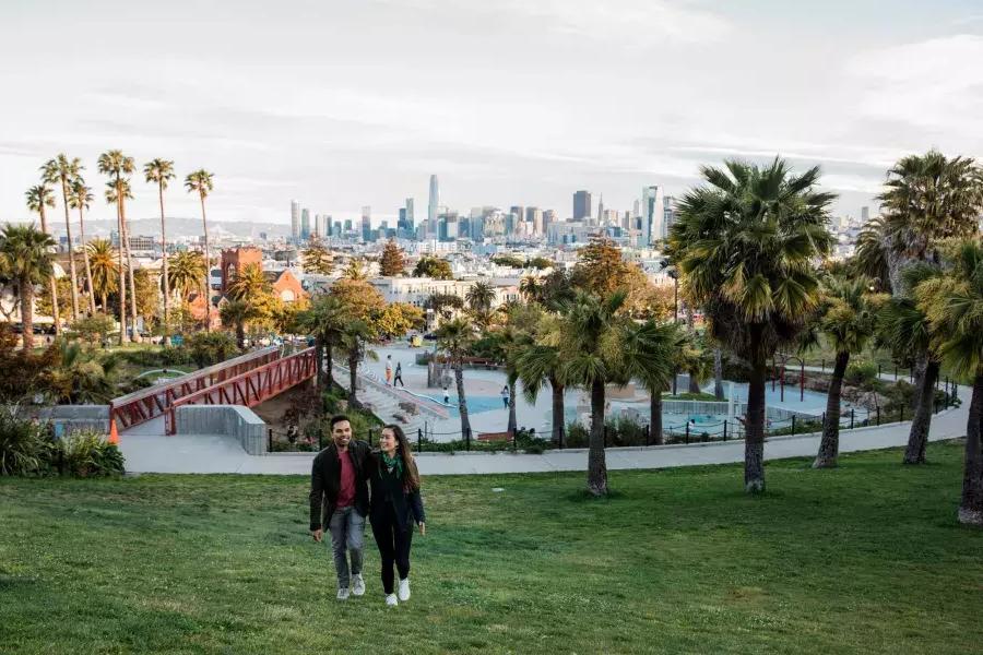 A couple walks toward the camera with Dolores Park and the 贝博体彩app Skyline behind them.