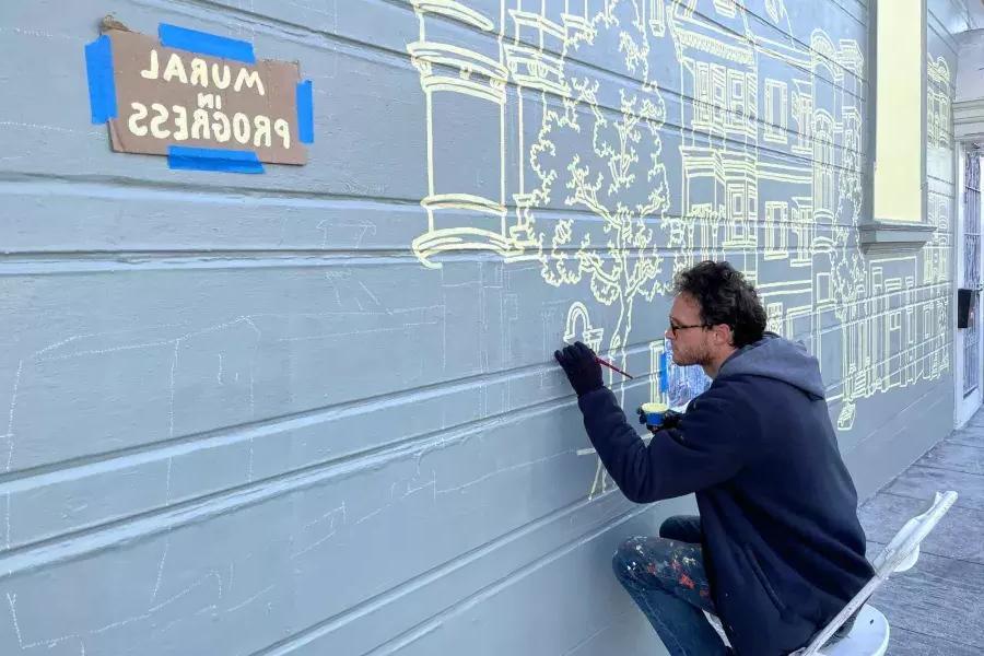 一名艺术家在Mission District一幢建筑物的一侧画壁画, 与一个标志在建筑上说“壁画正在进行中”. 加州贝博体彩app.