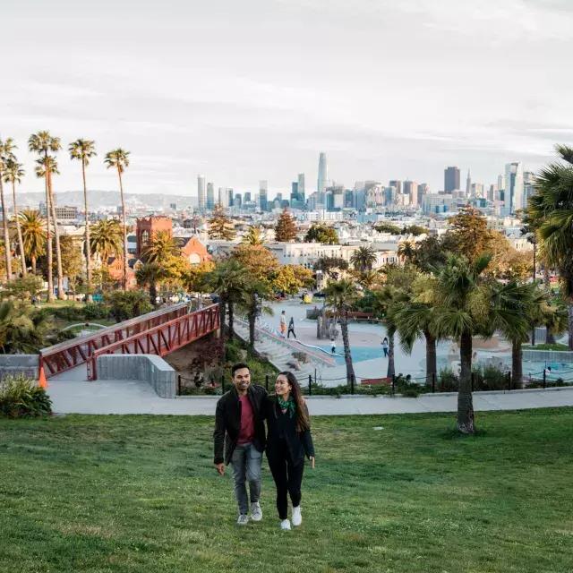 A couple walks toward the camera with Dolores Park 和 San Francisco Sky行 behind them.